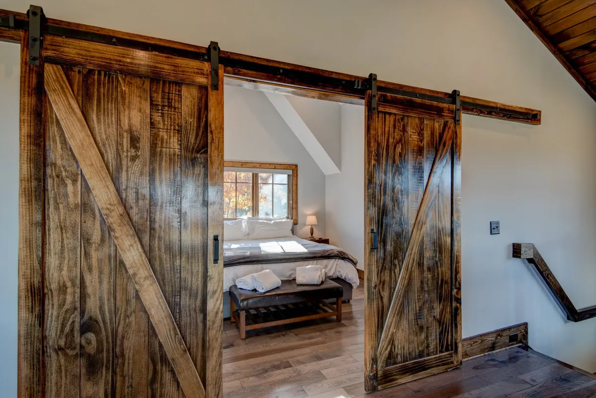 A serene master suite scene, featuring a bed with crisp white linens, a wooden bench, and large windows letting in abundant natural light. Sliding barn doors add rustic charm and privacy, hinting at the separation between the suite and a potential office nook.