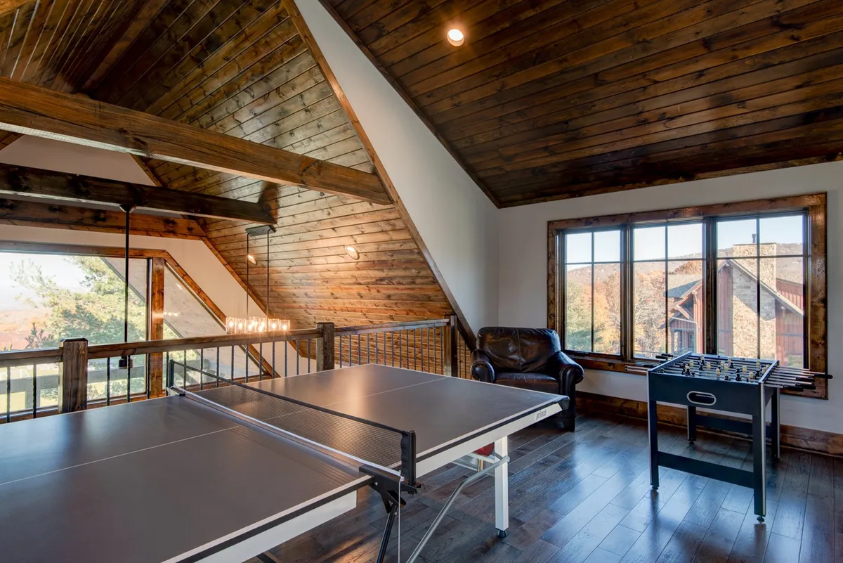 A bright, welcoming loft space featuring both a ping pong table and a foosball table set near a large window that frames scenic views. Soft, natural light fills the room, and the setup encourages friendly competition and group fun.