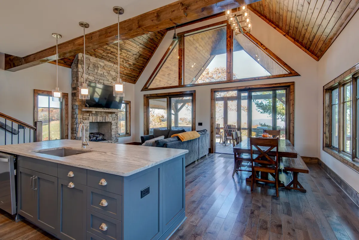A welcoming interior featuring a stone fireplace, a generously sized kitchen island with granite countertops, stainless steel appliances, and ample dining space. Large windows bring in natural light and frame the stunning outdoors, while wooden beams add a touch of rustic charm.