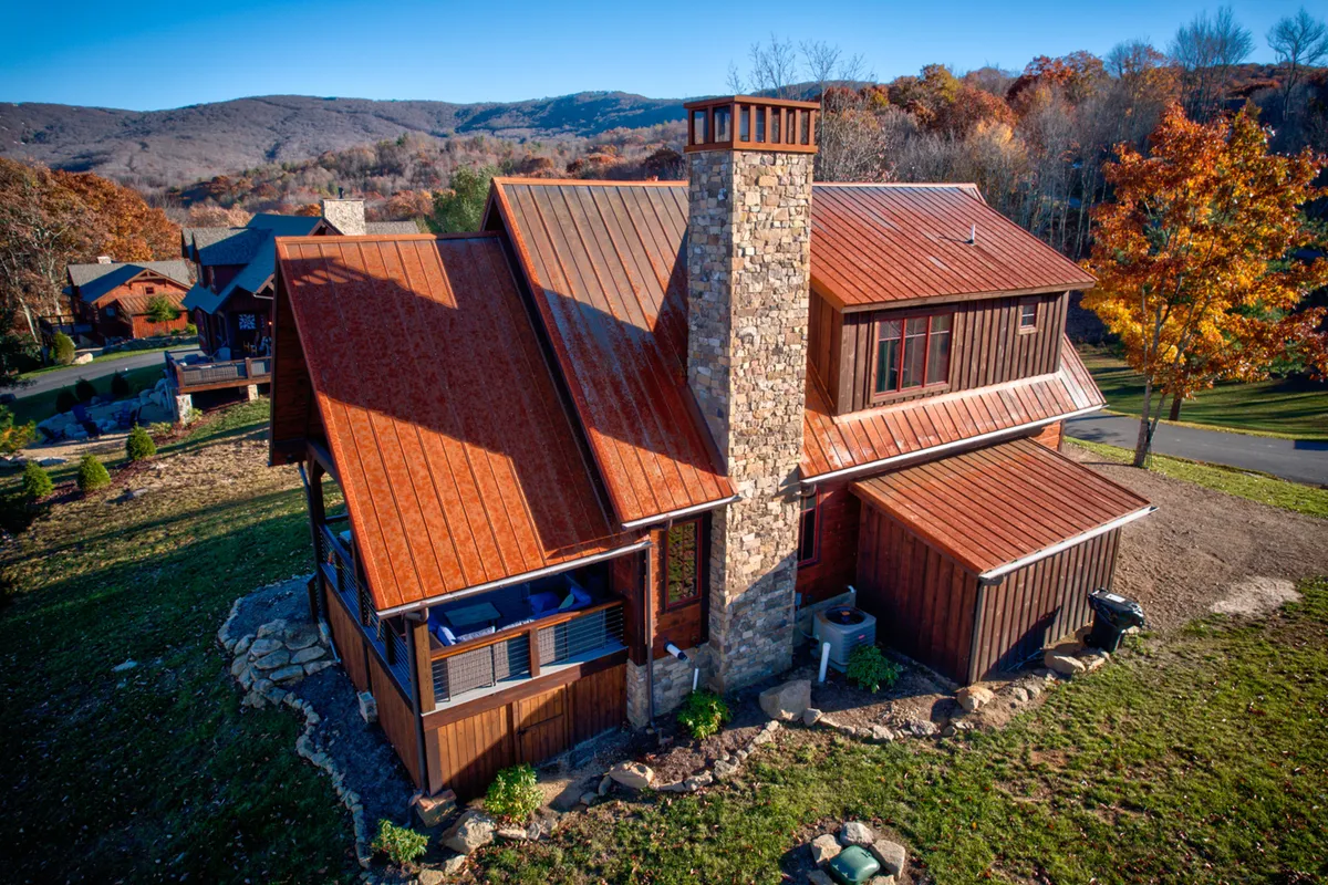 A charming exterior view of the property featuring a stone chimney, warm wooden siding, a modern metal roof, and a beautifully landscaped yard. The inviting front porch overlooks mountain vistas, creating a tranquil setting for morning coffee or sunset toasts.