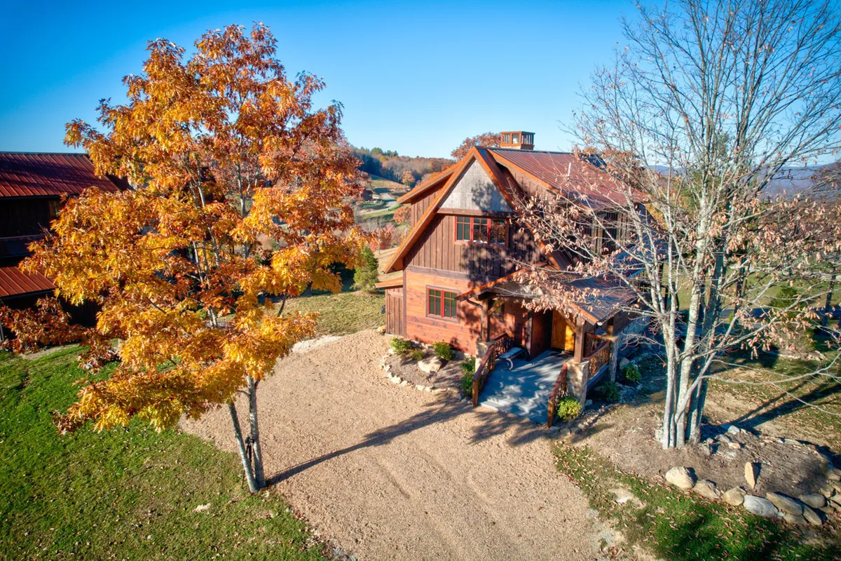 The image showcases a rustic wooden house nestled among vibrant autumn trees, with a gravel driveway leading up to a spacious covered porch. The inviting exterior is bathed in warm light, hinting at the cozy, welcoming atmosphere inside. The porch offers views of the changing mountain landscape, perfect for sunset watching or relaxing with loved ones.