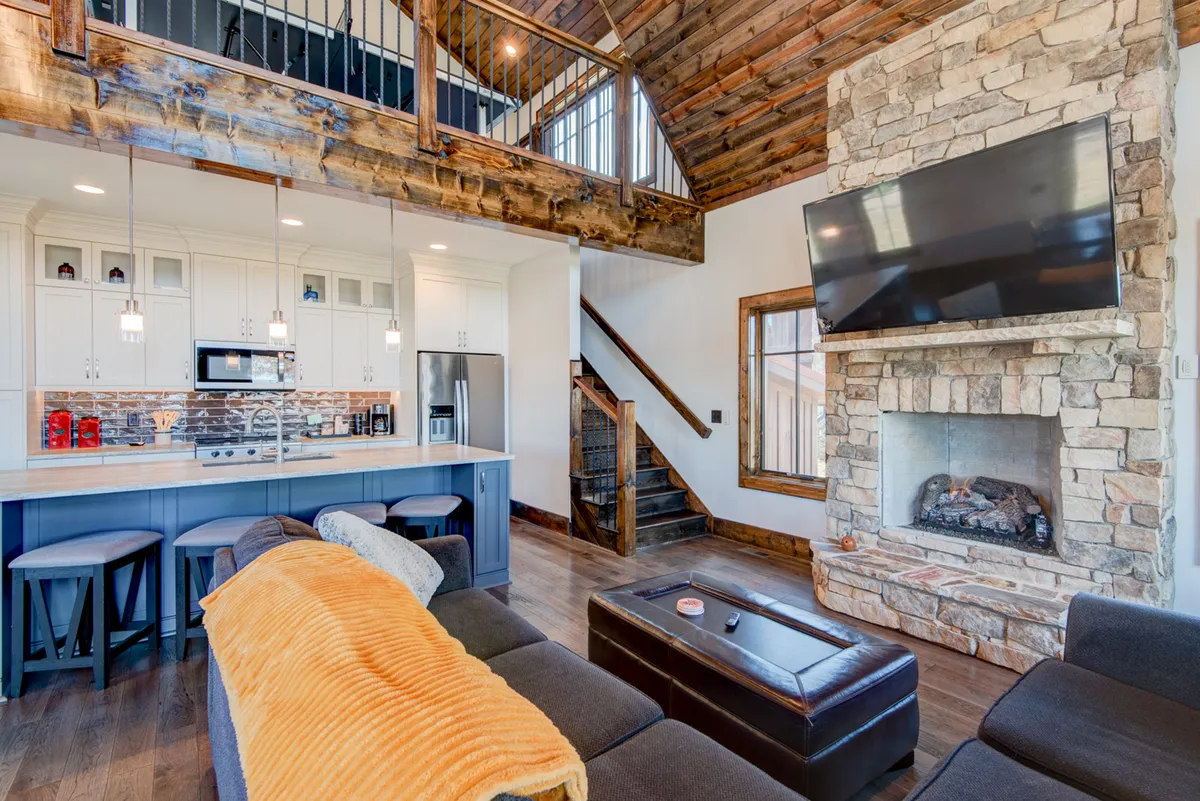 A warm, open great room with soaring cathedral ceilings, a striking Tennessee fieldstone fireplace, exposed wooden beams, and large windows that flood the space with mountain light. The kitchen island sits nearby, topped with granite counters and surrounded by bar seating, seamlessly connecting the living and cooking areas.
