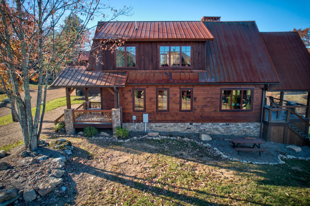 A rustic wooden facade with a metal roof and large windows sits nestled in lush greenery. The scene suggests ample space for outdoor family activities, gatherings, and relaxation under the open sky.