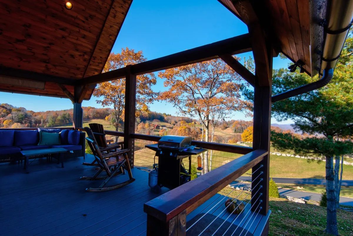A spacious covered porch with a comfortable couch, a classic rocking chair, and a grill, all oriented to showcase tranquil views of rolling hills and treetops. The outdoor seating area makes the most of the mountain scenery, inviting guests to relax, entertain, or simply watch the sunset.