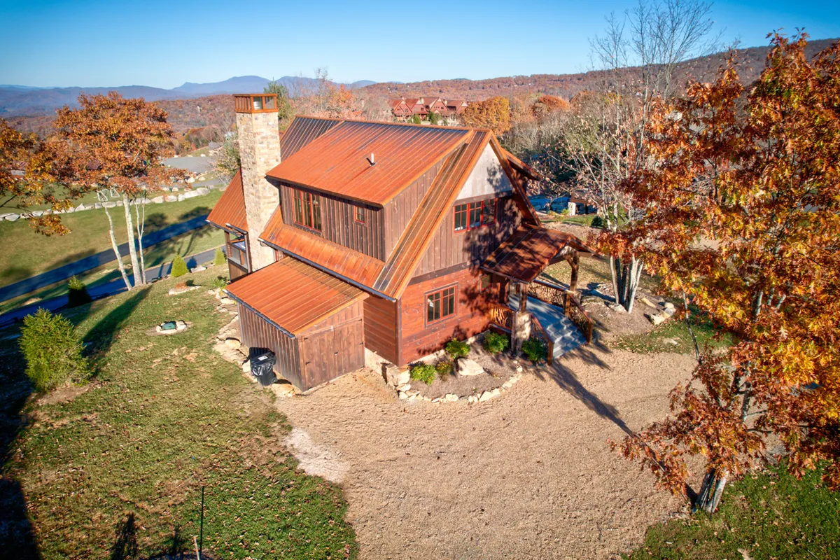 The rustic wooden cabin exterior is complemented by a vibrant red metal roof and framed by colorful autumn foliage. The surrounding gardens and mountain backdrop evoke feelings of seclusion and natural beauty, ideal for outdoor strolls and relaxation.