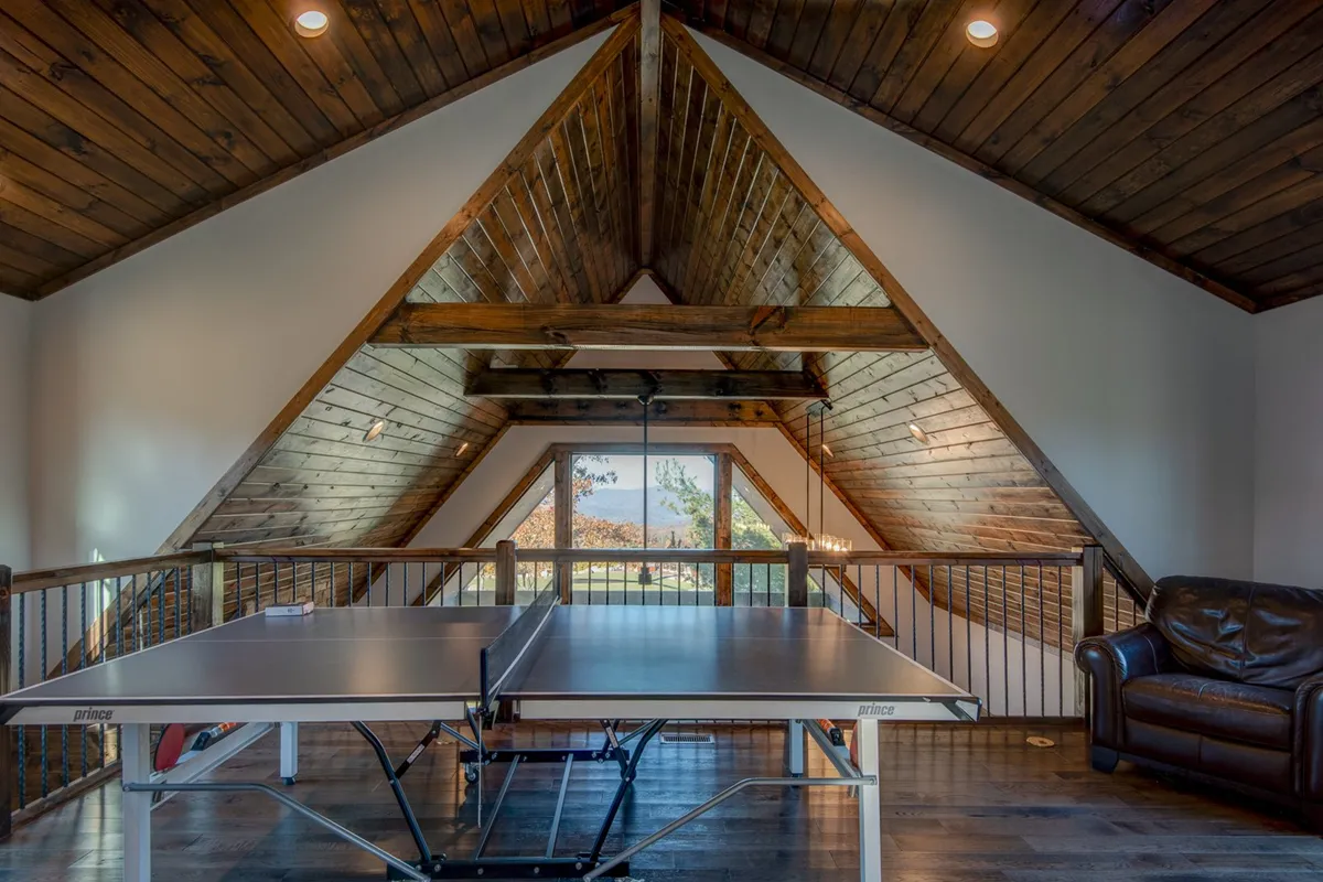 An interior recreation room featuring a ping pong table, large windows with scenic views, wooden beams, and a leather couch. The space is filled with natural light, suggesting a playful and relaxed mood perfect for friendly tournaments and gatherings with friends or family.