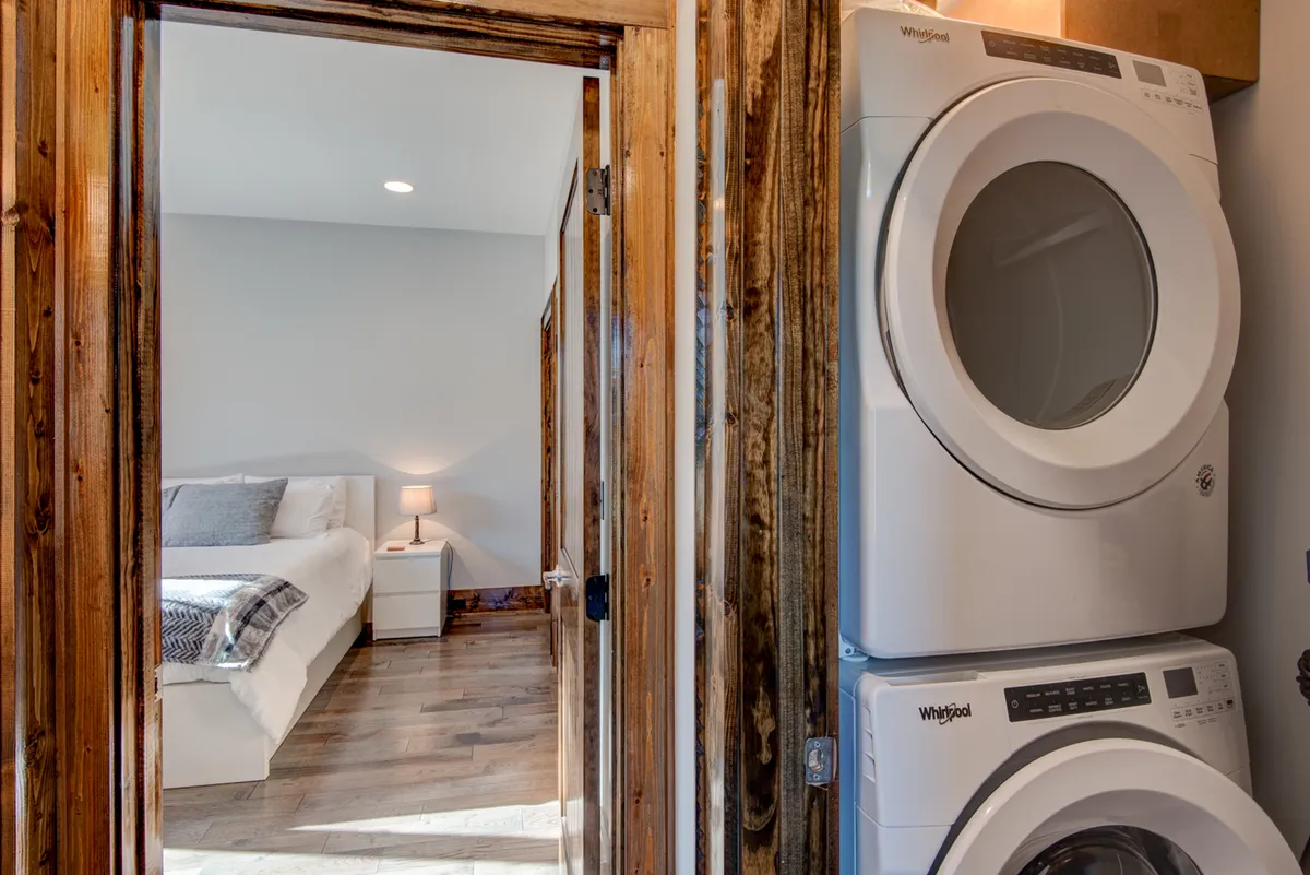 A functional space combining a laundry area with a cozy bedroom corner. The washer and dryer are neatly placed, while a nightstand and lamp beside the bed offer a blend of utility and comfort.
