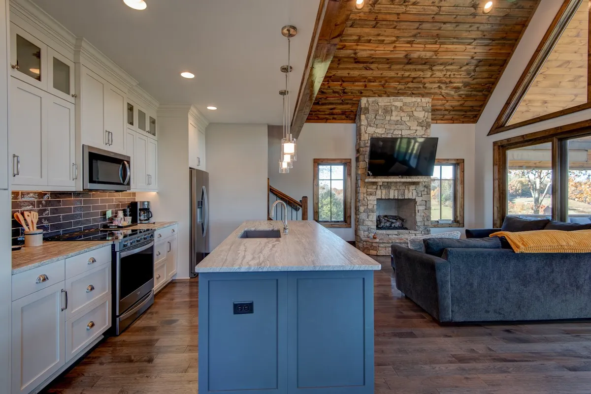 An expansive, light-filled great room with a comfortable sofa facing a rustic stone fireplace. The open layout reveals the adjacent kitchen, complete with a large island countertop and bar seating, all set against the backdrop of large windows that capture the scenic mountain views.