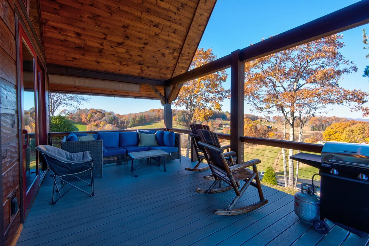 This image captures a spacious wooden deck adorned with inviting rocking chairs, a blue seating area, grill, and the vibrant backdrop of autumn trees. The setting is tranquil and radiates the allure of outdoor relaxation, social gatherings, and serene moments while highlighting both seating and dining possibilities under the cover of nature.